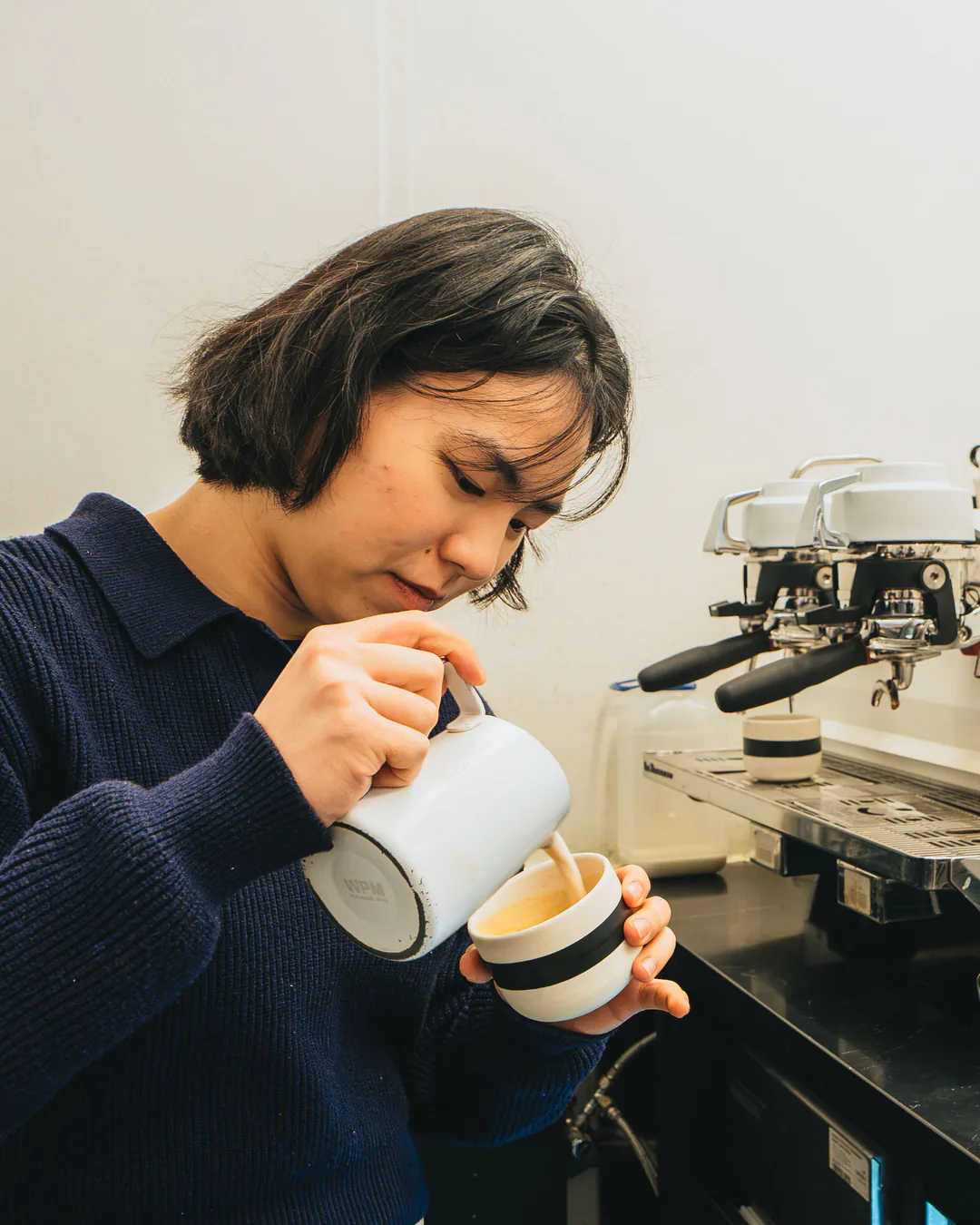 Barista gießt Milch aus einer weißen Kanne in eine Tasse, daneben Siebträgermaschine und Kaffeemühle