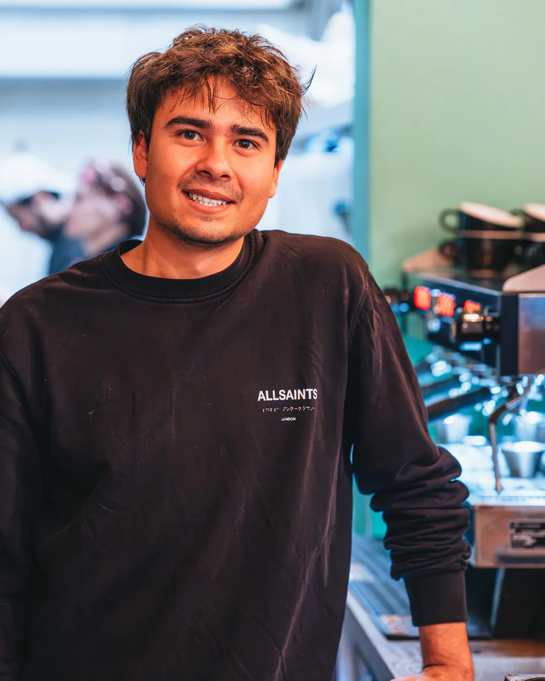 Ein junger Mitarbeiter im schwarzen Sweatshirt lehnt am Tresen und lächelt in die Kamera, rechts die Espressomaschine