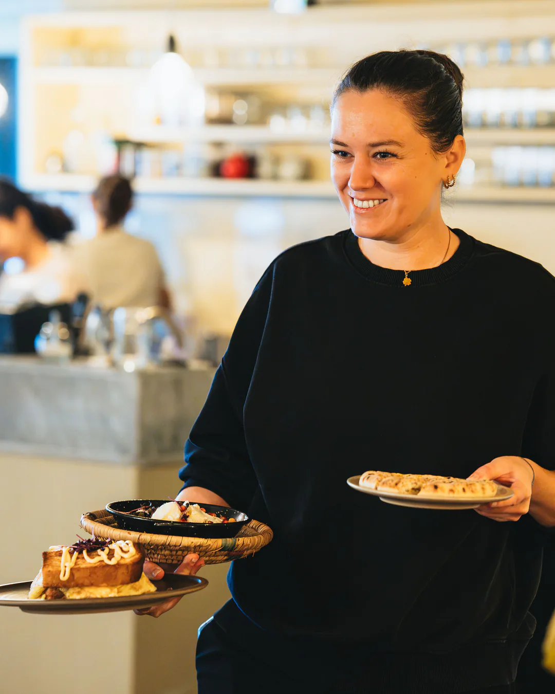 Lächelnde Servicekraft im schwarzen Sweatshirt trägt drei Teller mit Speisen, hinten der Tresen