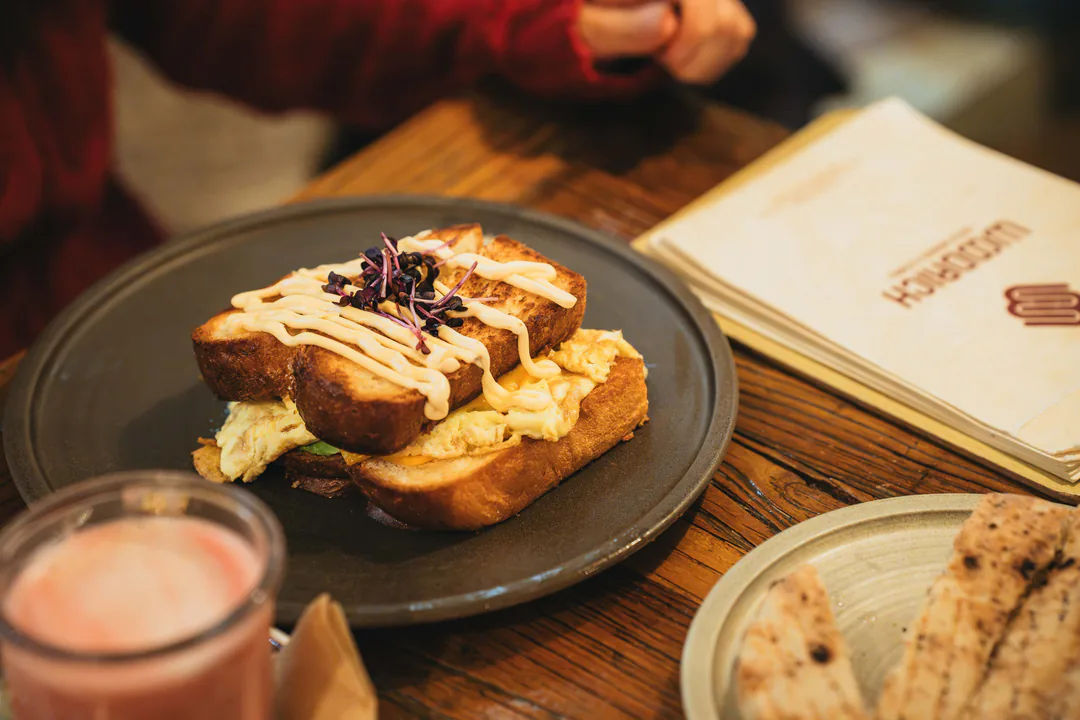 Getoastetes Sandwich mit Rührei auf einem Holztisch, daneben eine Woodrich-Speisekarte, ein rosa Getränk und Fladenbrot